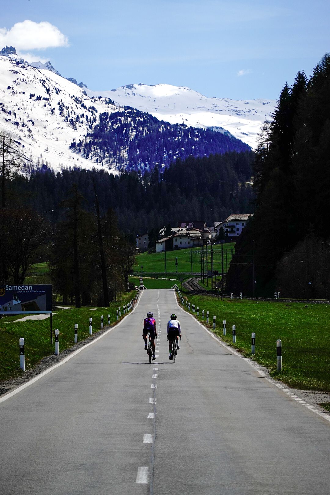 Two riders on a straight road towards snow-covered mountains near Samedan